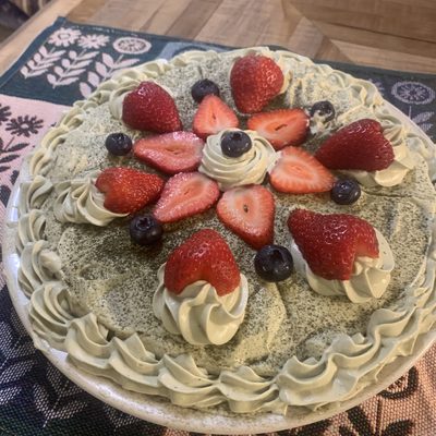 A matcha cream cake photographed from above, ringed with piped rosettes and topped with halved strawberries and blueberries arranged in a radiating pattern