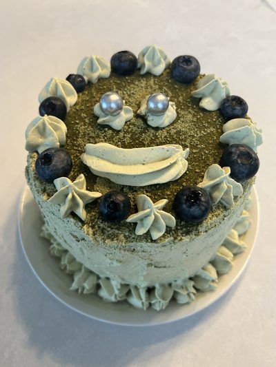 A small matcha layer cake dusted with green tea powder, decorated with a piped smiling face and a border of blueberries and pale green rosettes