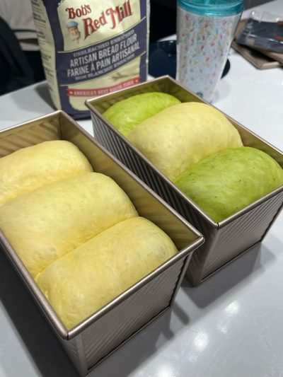 Two loaf pans of proofed milk bread dough, one plain and one tinted pandan green, beside a bag of bread flour