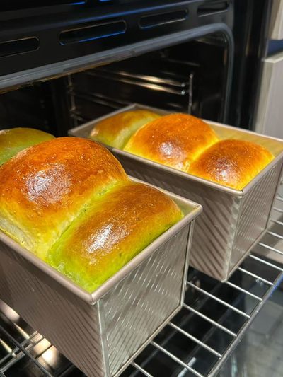 Two loaf pans of milk bread browning inside an oven, one plain and one tinted pale green with pandan