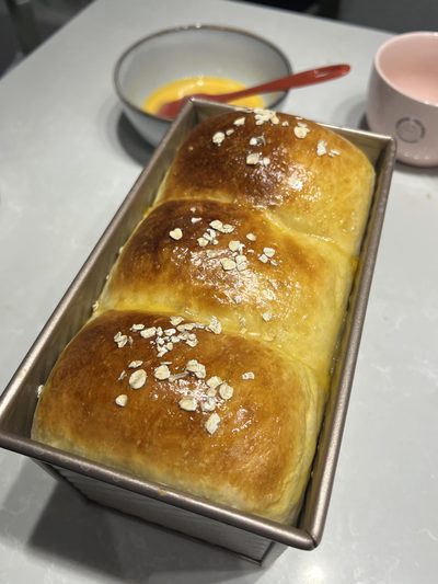 A three-lobed milk bread loaf resting in its pan, glazed and scattered with rolled oats, next to a bowl of leftover egg wash