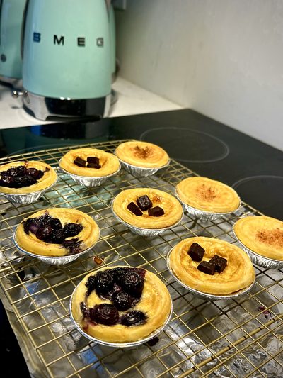 Egg tarts and blueberry tarts in foil cases cooling on a wire rack, with a mint green electric kettle behind them