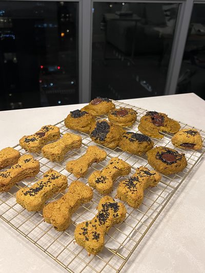 Bone-shaped pumpkin dog biscuits topped with black sesame seeds, cooling on a wire rack beside a batch of cookies