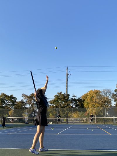 A tennis player mid-serve on an outdoor blue hard court, racket raised and ball in the air against a clear sky, with autumn trees behind the fence