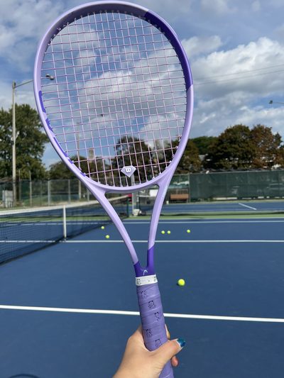 A lavender tennis racket held up against a blue hard court, balls scattered across the far side and cloud overhead