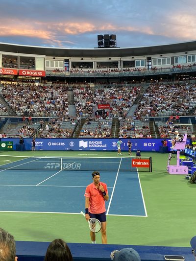 An ATP Tour match on a stadium court at sunset, pink clouds above a full crowd, a player standing at the baseline