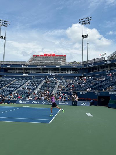 A tennis player following through on a shot on a stadium court under a blue sky with scattered cloud, spectators in the stands behind