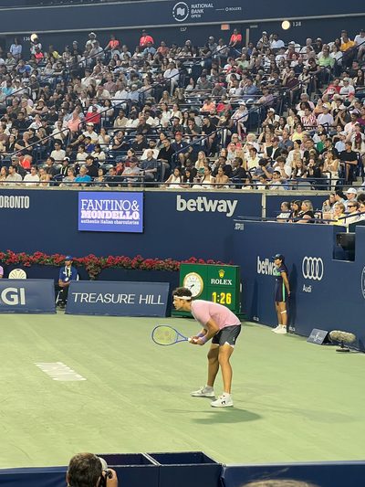 A player crouched low to receive serve on a stadium court, the stands packed behind and a tournament clock at the baseline