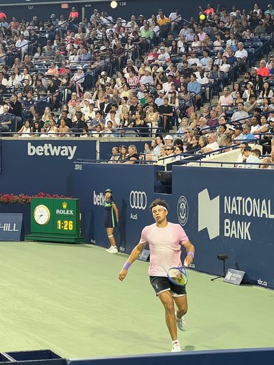 A player sprinting along the baseline of a stadium court to reach a ball, sponsor boards and a full crowd behind