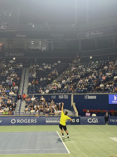 A tennis player tossing the ball to serve on a floodlit stadium court at night, the air hazy with light