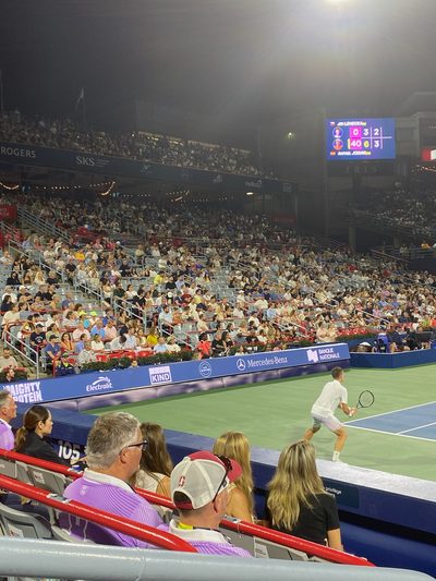 A night tennis match seen from the spectator stands, the scoreboard lit up beyond the court and a player about to hit a forehand