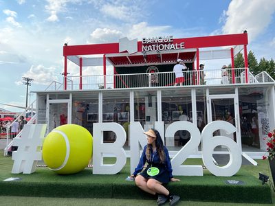 A person sitting in front of oversized white letters spelling a tournament hashtag next to a giant tennis ball sculpture on a sunny day