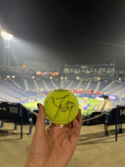 A hand holding up a signed tennis ball in front of an empty floodlit stadium late at night