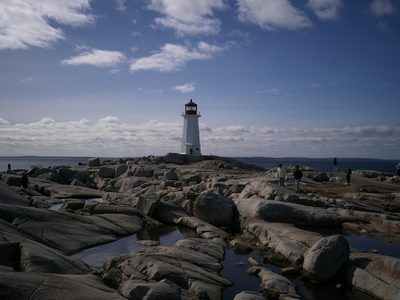 A white lighthouse standing on wave-smoothed granite at Peggy's Cove, Nova Scotia, under a wide sky of scattered cloud with tide pools in the foreground