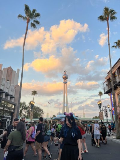 A person in Mickey Mouse ears walking down a theme park boulevard at sunset, palm trees and a tall art deco tower catching the last light